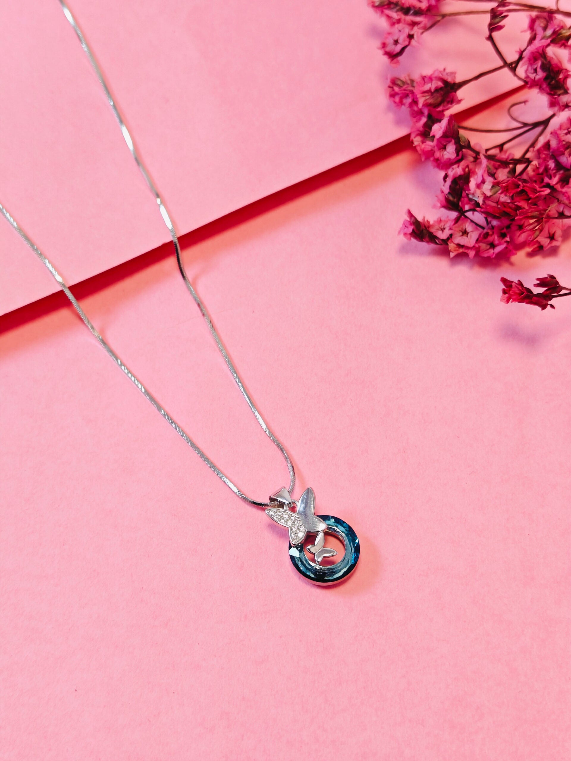 Silver necklace with a blue and clear pendant on a pink background with pink flowers.